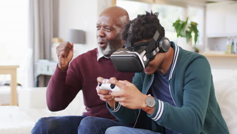 happy african american son using vr headset and gamepad with senior father at home, slow motion