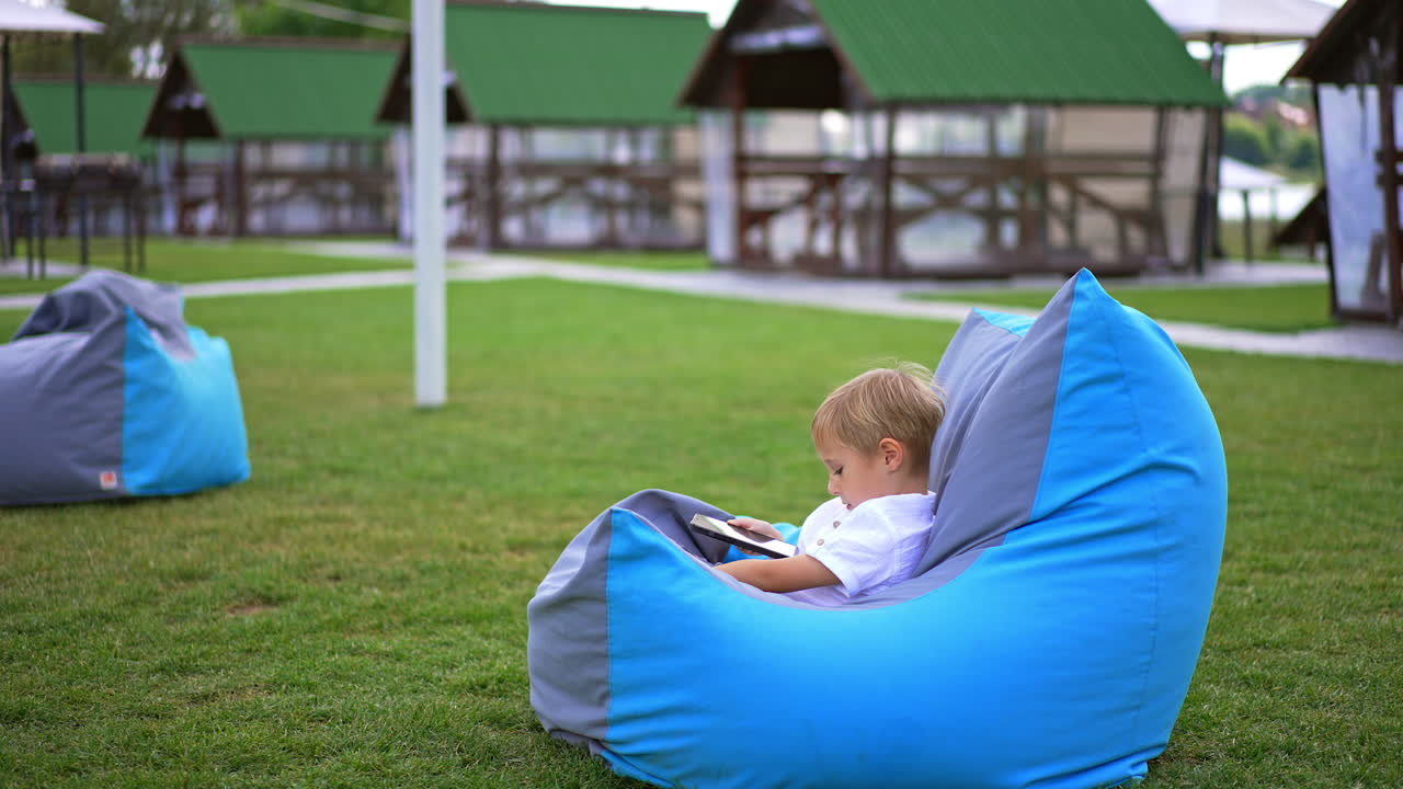 Little six-year-old blond boy sitting comfortably in bean bag chair with phone in his hands. Child resting outdoors with gadgets. Blurred backdrop.
