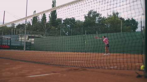 Imágenes-Comerciales-En-Cámara-Lenta-De-La-Práctica-De-Tenis-A-Través-De-La-Red-De-La-Cancha-De-Tenis.-Vista-Directa-De-Una-Atleta-Jugando-Al-Tenis.-Un-Deportista-Adolescente-Golpea-La-Pelota-Durante-El-Entrenamiento-Deportivo