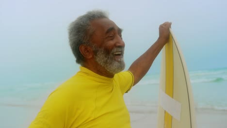 vista frontal de un hombre afroamericano senior activo y feliz con una tabla de surf de pie en la playa 4k