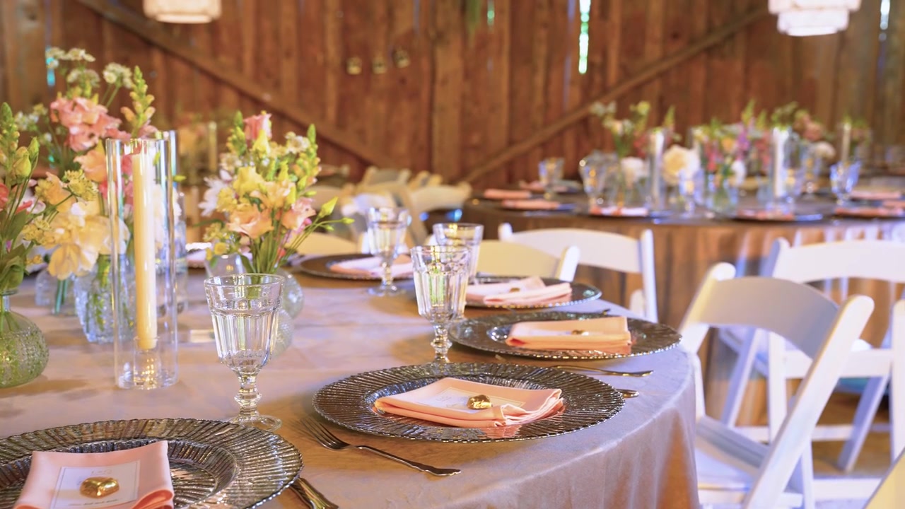 Charming rustic barn wedding reception setup with pastel floral centerpieces, glassware, metallic chargers, and peach napkins on round tables in warm, natural lighting. Stylish and inviting.