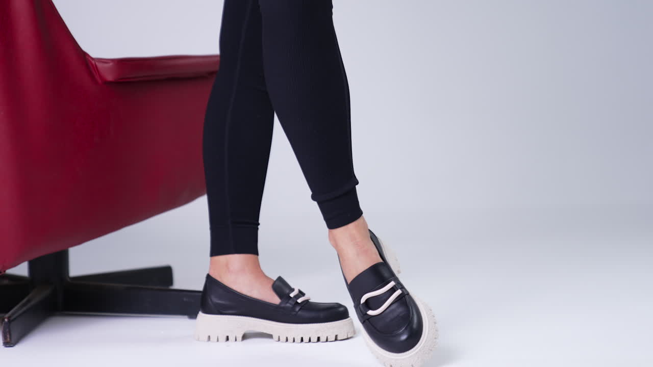 Unrecognized female in black pants wears modern shoes on white soles. Woman standing beside red chair demonstrating stylish footwear. White backdrop.