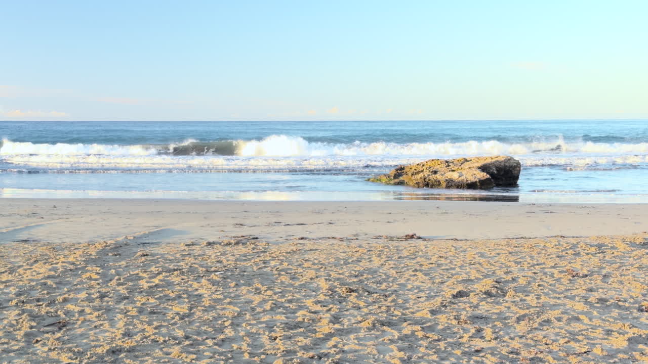 Early morning view of a quiet sandy beach with waves breaking near a coastal rock. Bright natural light and gentle ocean motion create a peaceful and scenic Mediterranean seascape