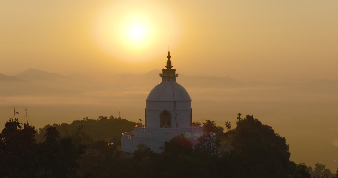 Aerial gliding shot towards the Peace Pagoda World Peace Stupa in Pokhara Nepal with golden sunrise light and warm sky illuminating the surrounding landscape.