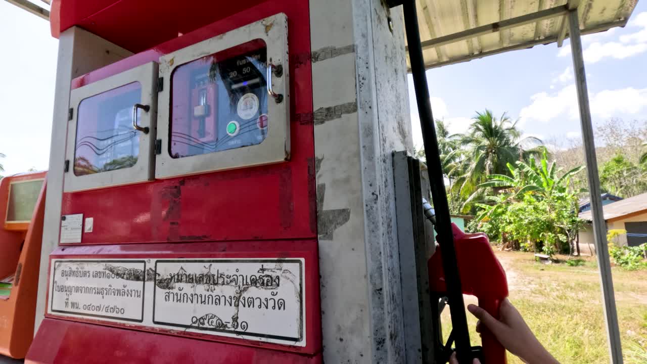 Person operates retro fuel pump outdoors in bright daylight, rural Phuket, handheld camera movement