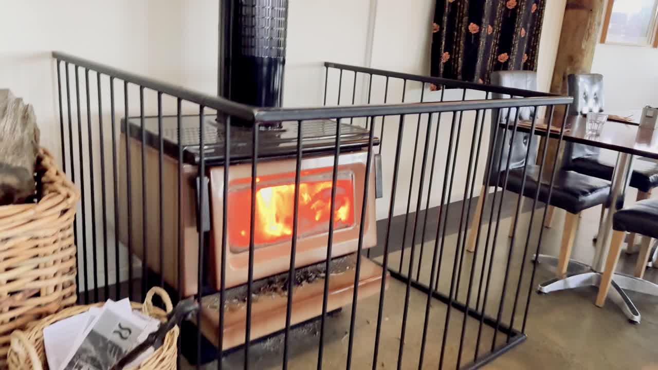 A wood-burning stove enclosed by a safety gate radiates warmth in a cafe or restaurant setting, with natural daylight and steady handheld camera movement