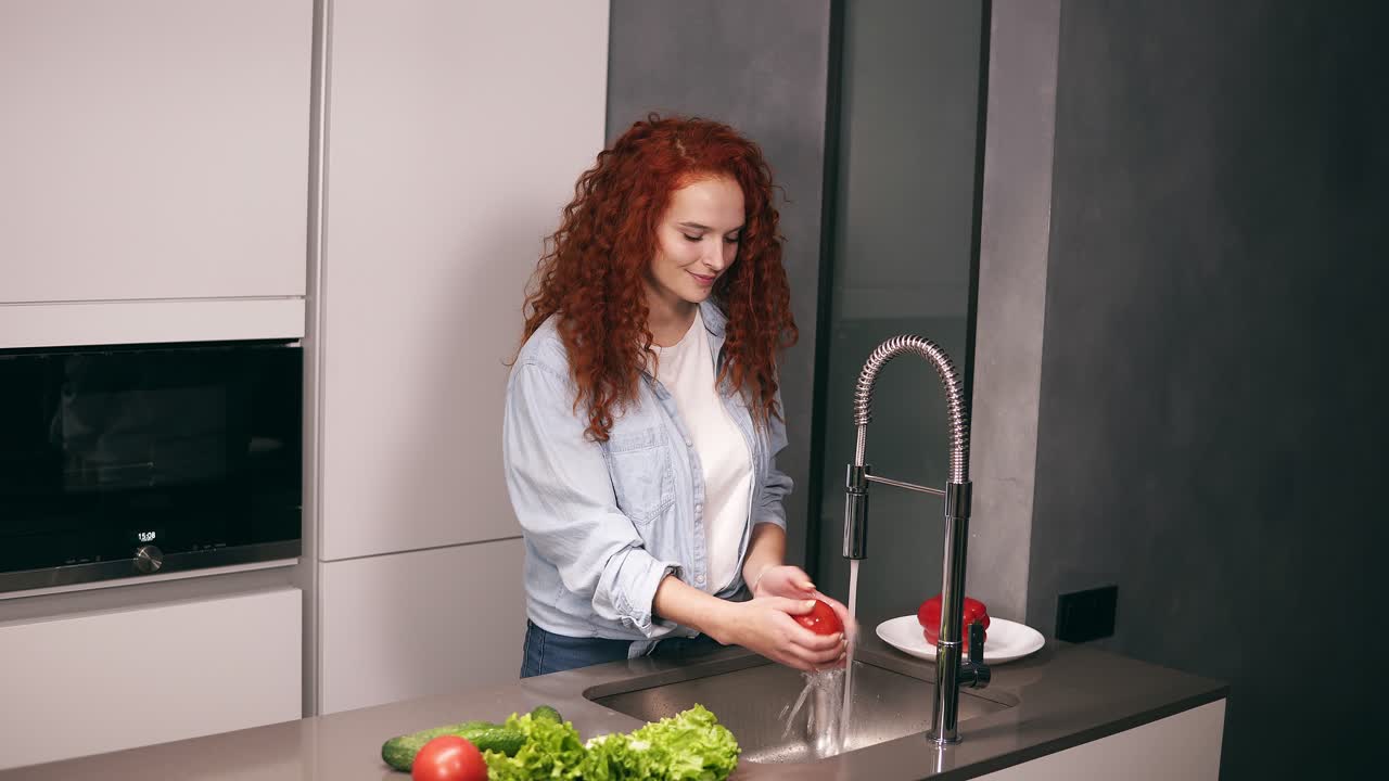 Affascinante donna dai capelli rossi lava peperoni rossi, cetrioli e pomodori in cucina e li mette su un piatto. Concetto di cibo fatto in casa.
