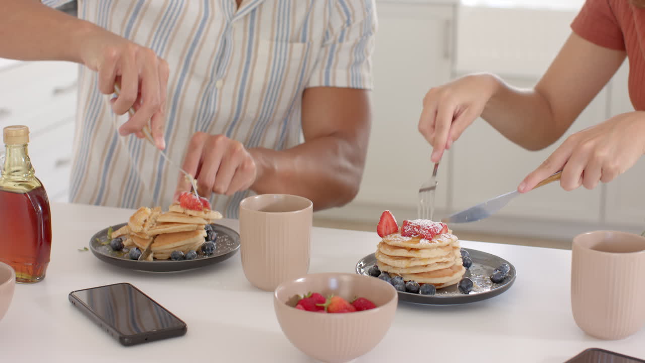 Eating breakfast with pancakes and strawberries, diverse couple smiling and chatting at home