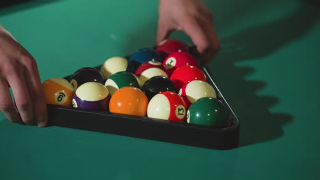 close-up view of guy's hand in white shirt and gray trousers carefully rolling colorful billiard balls in triangle rack on green pool table, ensuring precise alignment for a perfect break