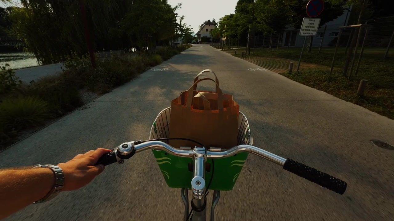 POV view of a person riding a city bike beside a river, with a reusable shopping bag in the front basket and warm evening light along the path.