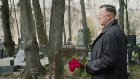 side view of man in black raincoat and suit holding roses while walking in a graveyard