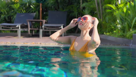 asian girl standing in a swimming pool, wearing a yellow bikini and sunglasses correcting her wet hair and looking into the camera afterwards