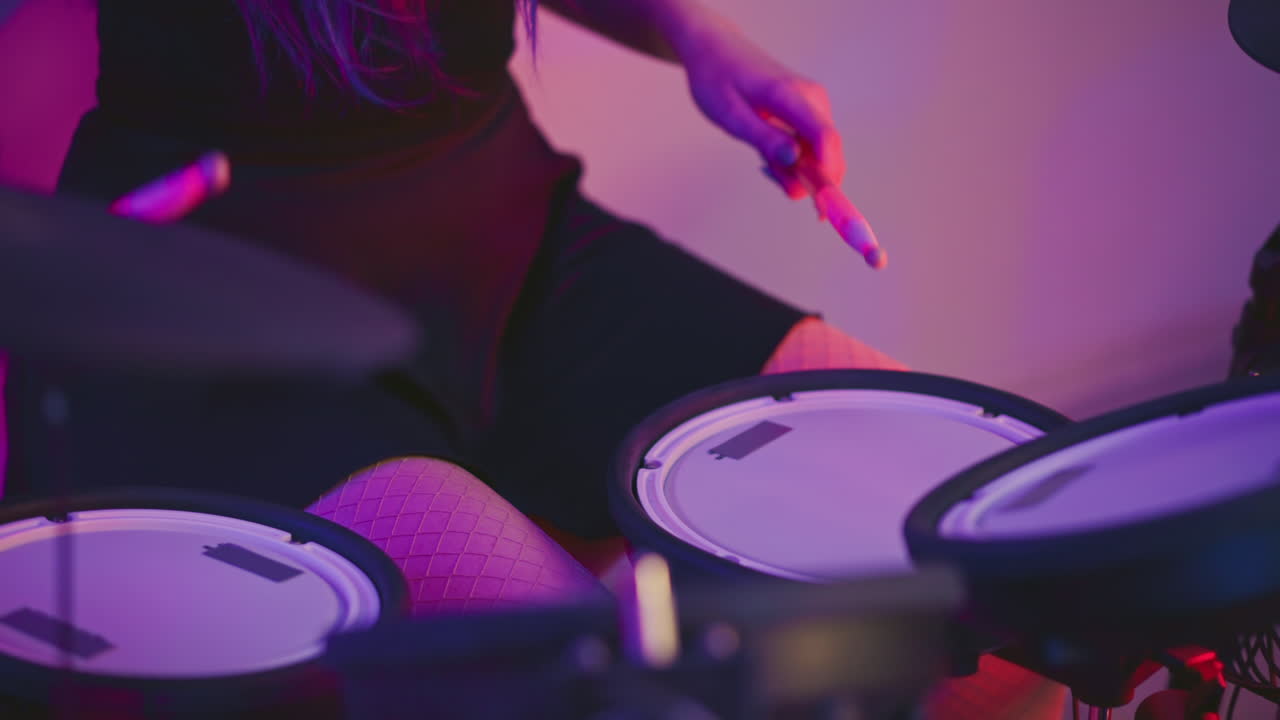 Partial view of girl wearing fishnet pantyhose seated at electronic drum set, casually holding drumstick while practicing under soft purple and pink lighting inside studio with relaxed vibe