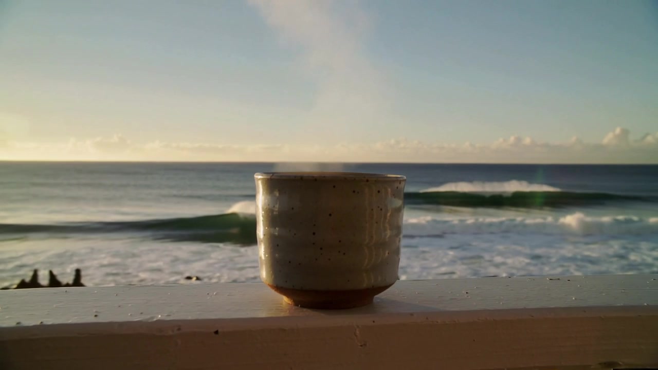 Telecamera che riprende una tazza sulla ringhiera di un ponte, mostrando il bagliore dell'alba e la mareggiata che spinge le onde.