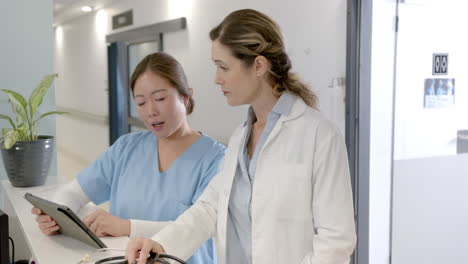 focused diverse female doctors using tablet and discussing at hospital reception desk, slow motion