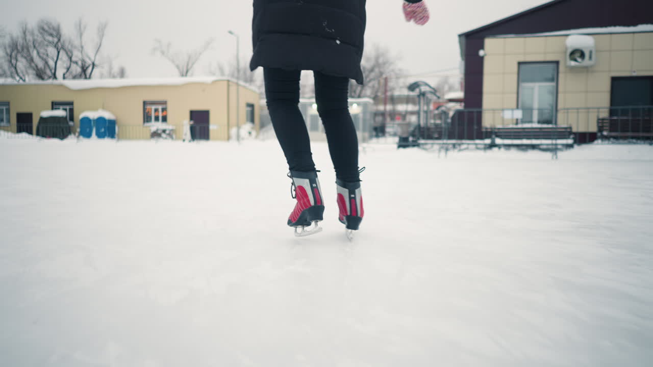 Lady in black coat skating outdoors on snowy winter day, captured from behind focusing on red skates gliding across frozen surface with snow-covered surroundings in soft blurred background