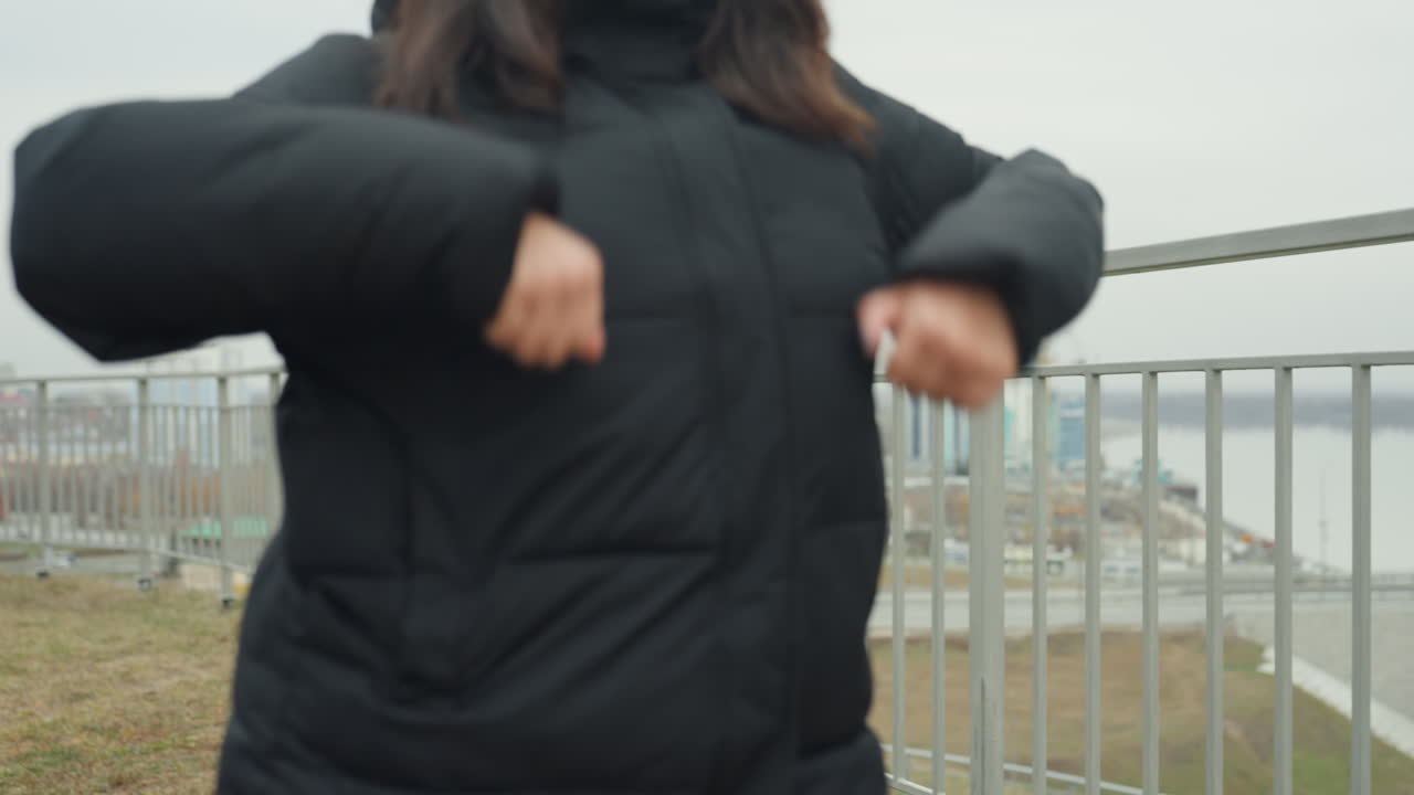 Smiling caucasian woman dancing on rooftop, knit beanie and puffer jacket, arm gestures and light choreography, joyful expression, city skyline backdrop, spontaneous rehearsal vibe