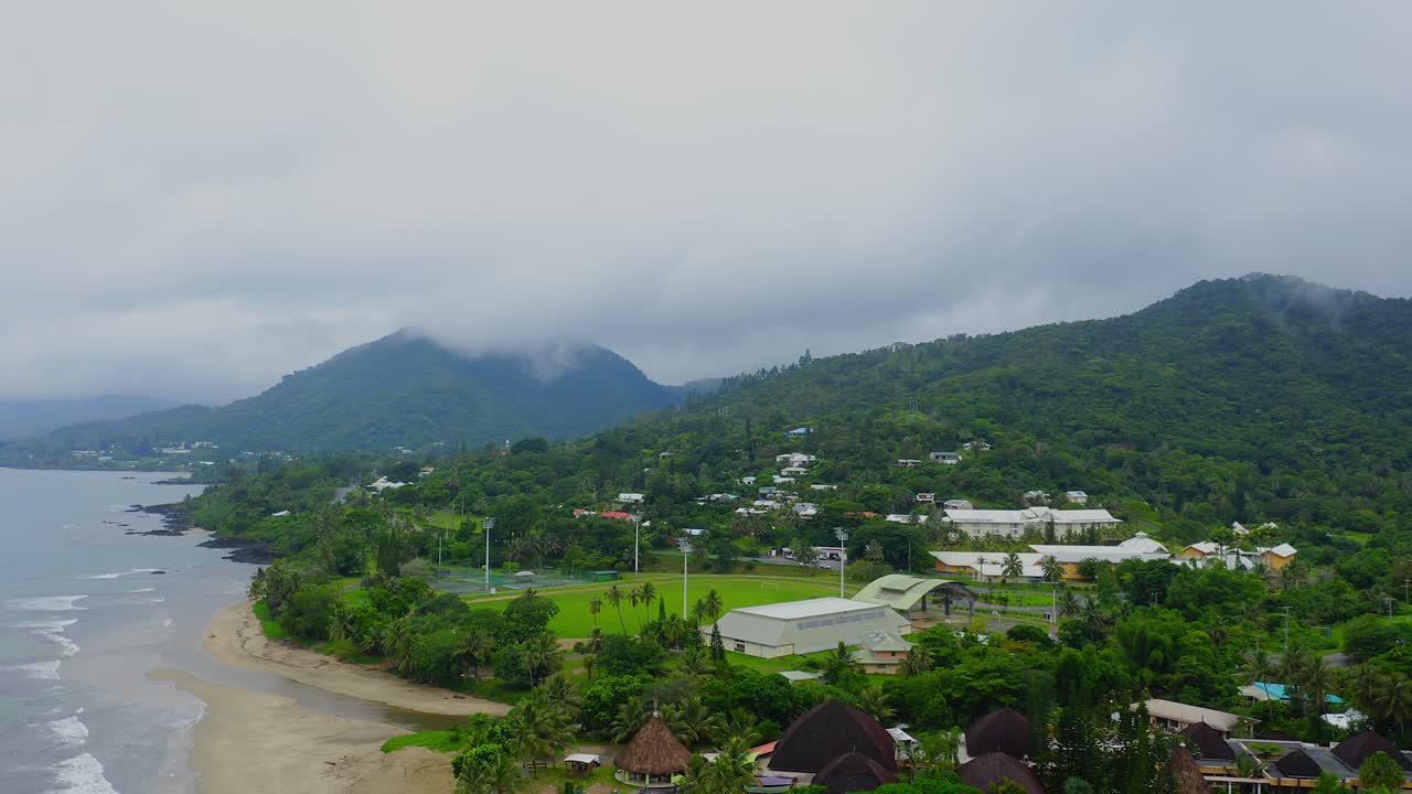 imágenes aéreas que se mueven hacia atrás sobre un paisaje urbano tropical de nueva caledonia con exuberantes colinas boscosas y una playa costera en un día lluvioso