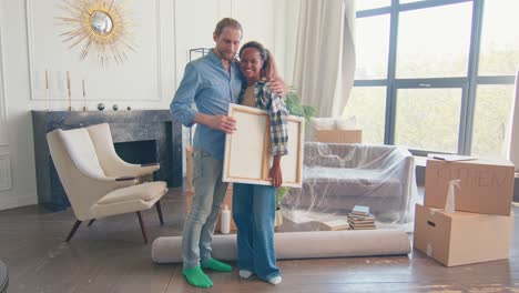 diverse lovers couple moving to home and holding painting stands in living room