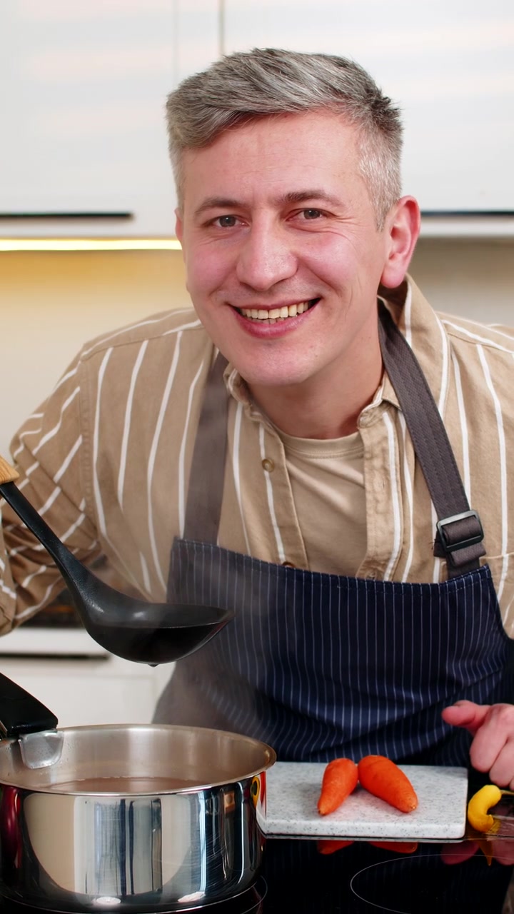 Mann mittleren Alters bereitet lächelnd gesunde Diätsuppe in der Küche zu, während er zu Hause eine hausgemachte Mahlzeit zubereitet