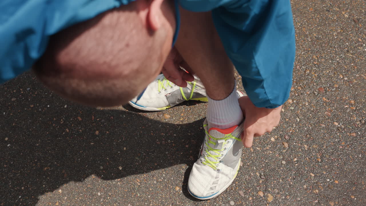 Primo piano dall'alto di un uomo bianco che allaccia una scarpa da football all'aperto, che mostra un giocatore calvo con una giacca blu che stringe una scarpa con lacci al neon sull'asfalto bagnato, mani e calzini concentrati, rituale pre-partita in condizioni di pioggia