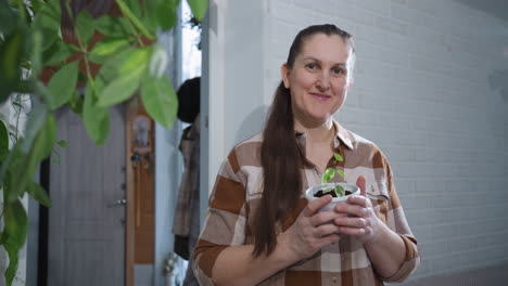 green thumbed caretaker holding plastic planter with variegated seedling, gently swaying body in motion while admiring new growth against indoor backdrop adorned with hanging pots and leafy greens