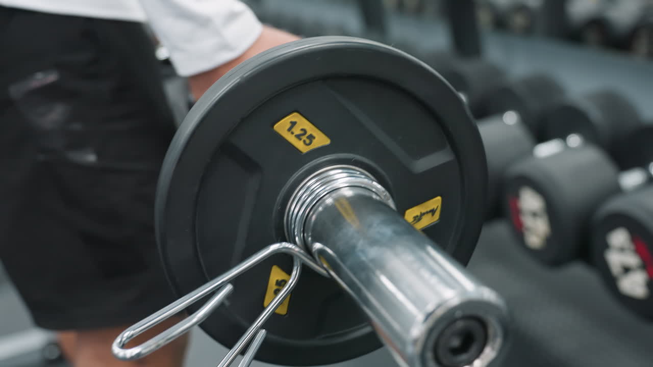 Close up of athlete gripping dumbbell performing strength exercise with blurred background showing rack and gym members on rubber flooring capturing workout intensity and muscle focus