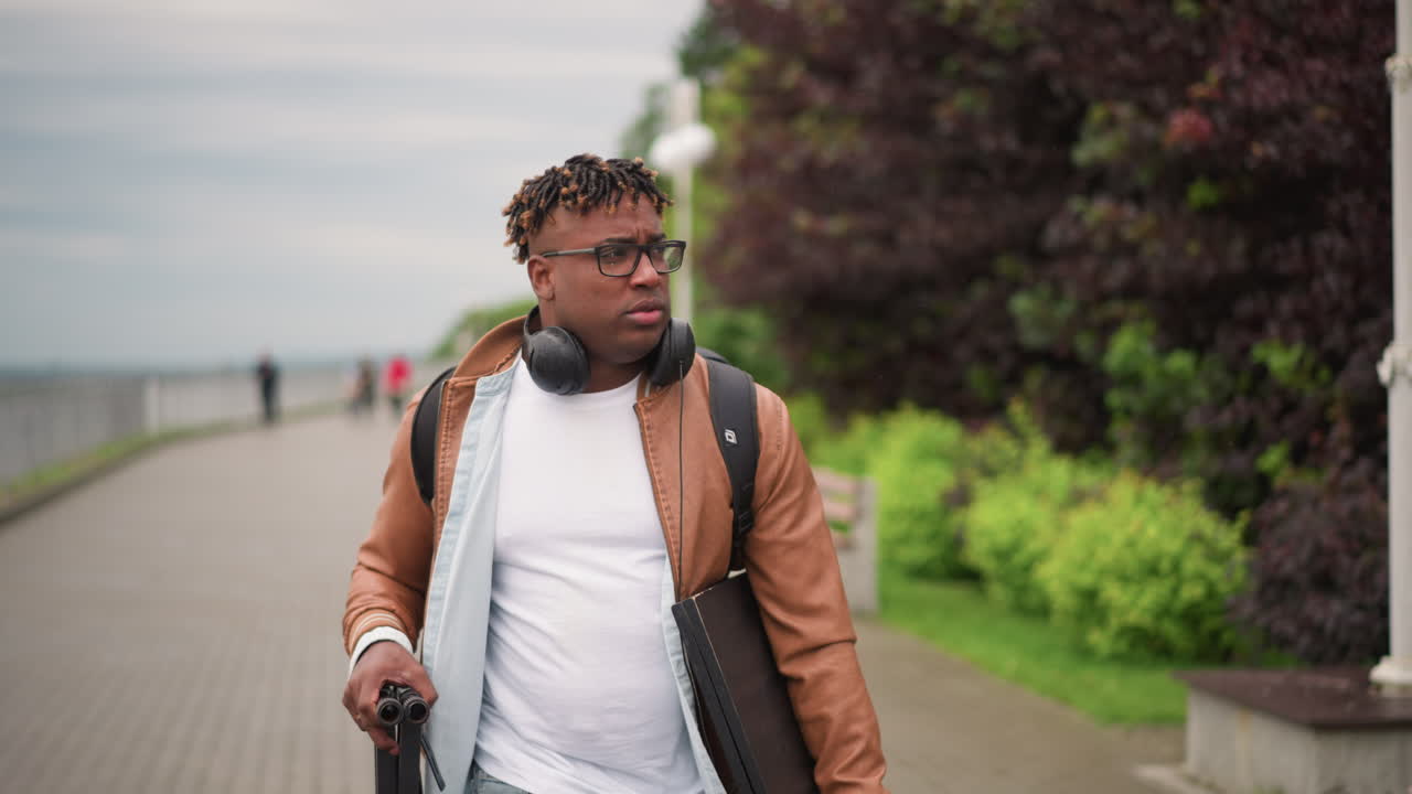 Black man walking coastal boardwalk, headphones, brown jacket and ...