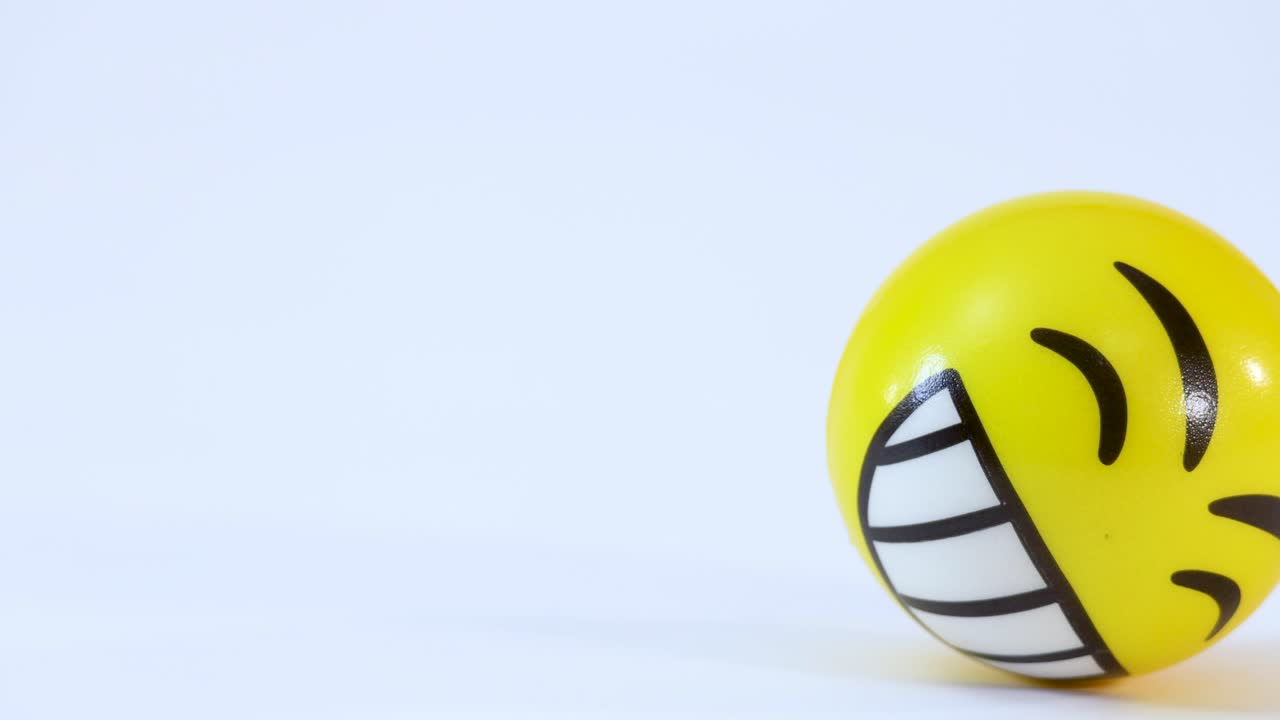 Bola stres emoji kuning bergulir di atas permukaan putih polos di bawah pencahayaan studio yang terang.