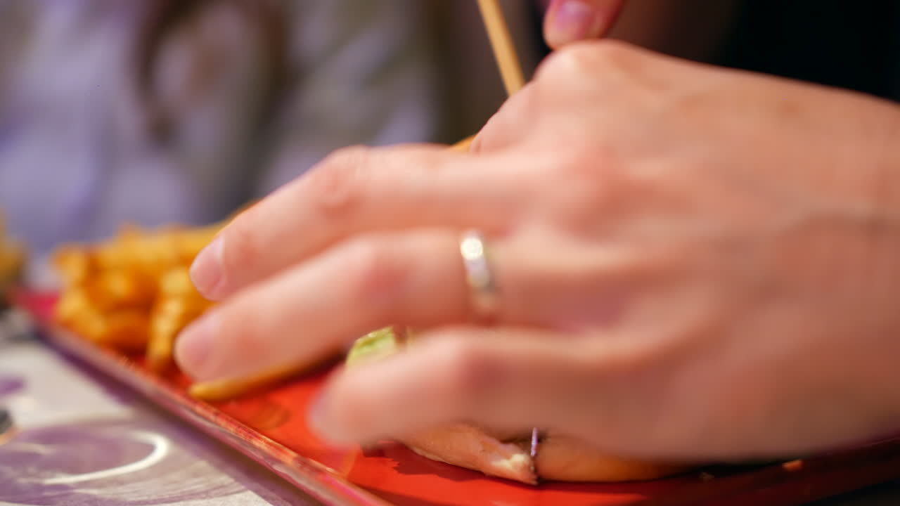A woman slicing and cutting a juicy cheeseburger topped with lettuce and tomato, served on a toasted bun. The burger sits on a rectangular plate next to a side of golden fries, ready to be enjoyed