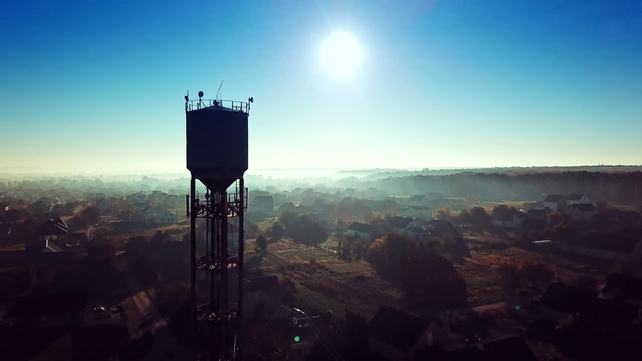 Flying over the village near the power station tower on a blue background sunrise. Countryside panorama in the morning dawn. Aerial view.