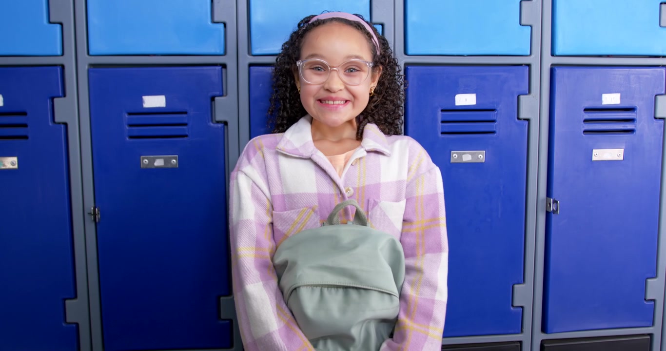 African American female teen facing camera holding sage backpack at lockers, softening and smiling