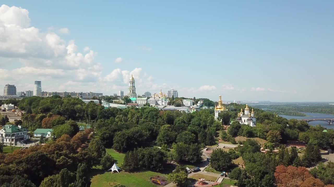 Ukraine,Kyiv,Pechersk Lavra,Drone footage zooms in to the Lavra's main buildings,towers on a sunny day with white clouds.Building Complex surrounded with green trees,Dnipro river, bridge, golden domes