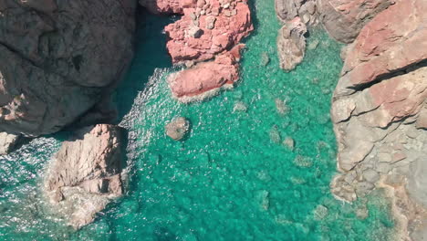 Vista-Superior-Hacia-Arriba-De-Las-Rocas-Rojas-Con-Olas-Rompientes-En-Un-Hermoso-Mar-Azul-Y-Agua-Tropical-En-Un-Día-Soleado-En-Cerdeña,-Italia---Toma-Aérea-De-Drones