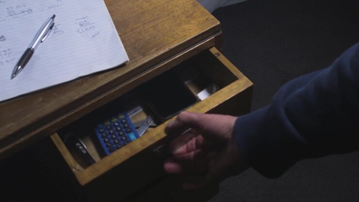 Mano adulta apre scorrendo un cassetto di scrivania in legno accanto a appunti di bilancio scritti a mano, rivelando uno smartphone, una calcolatrice, bollette e cancelleria.