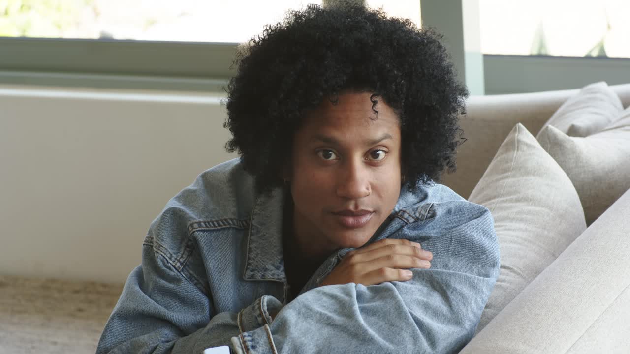 Reflecting, mid-adult African American man leaning on sofa at home shifting gaze, smiling at camera