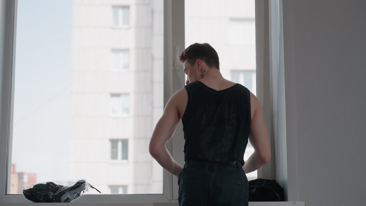 Fair skinned man in black tank top stands at bright window of modern high rise apartment, adjusts black jacket on shoulders then smooths fabric as cityscape blurs outside in soft daylight