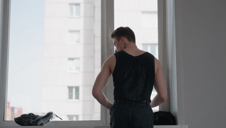 fair skinned man in black tank top stands at bright window of modern high rise apartment, adjusts black jacket on shoulders then smooths fabric as cityscape blurs outside in soft daylight