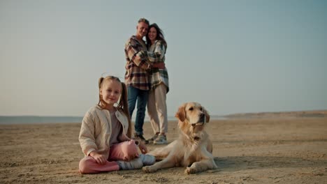 retrato de una feliz niña rubia en una chaqueta blanca y pantalones rosados que se sienta y se relaja y acaricia a su gran perro de color crema detrás de ella son sus padres que están abrazando y viendo con su niña mientras se relajan en una playa desierta en verano
