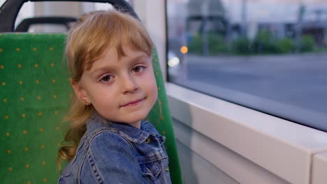 cheerful cute child girl passenger riding at public modern bus or tram transport, looking out window