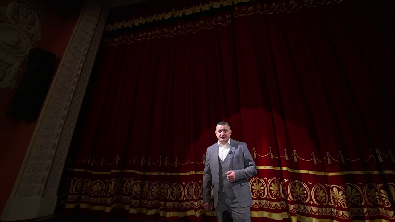 Actor with microphone announces performance before auditorium. Theater curtains background.