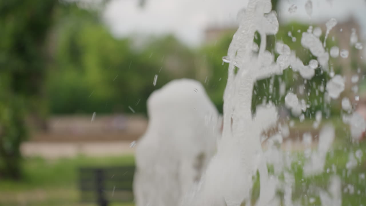 Water fountain flow tapering off while people seated on bench blur in background, summer park atmosphere with soft green trees and fading spray, dynamic droplets catching light