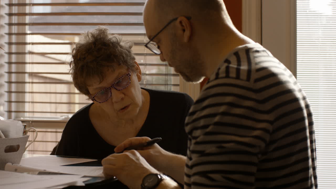 An elderly woman sits at a kitchen table with a middle-aged man. The man reviews and signs documents, while the woman looks over his shoulder. Both wear reading glasses. Warmly sun lit through window.