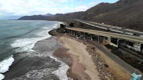 aerial drone shot orbiting around a concrete bridge on the beach in ventura with surfers in the ocean and rv campers next to the california 101 freeway along the coast