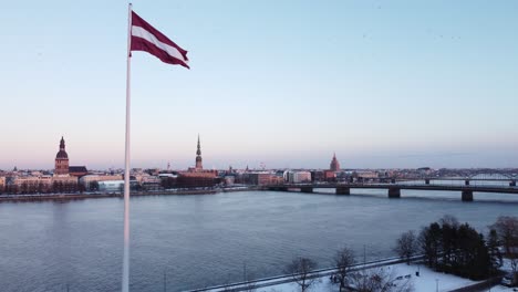 Malerische-Luftaufnahme-Der-Nach-Vorne-Fliegenden-Lettischen-Flagge-An-Der-Stange,-Die-Das-Stadtbild-Zeigt