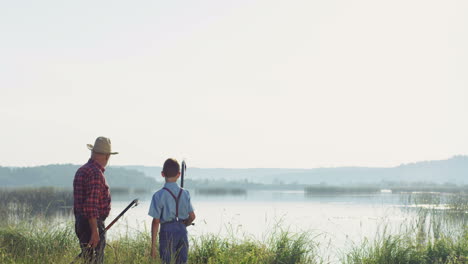 rear view of a grandfather and grandson standing and talking while choosing a place for angling on the lake