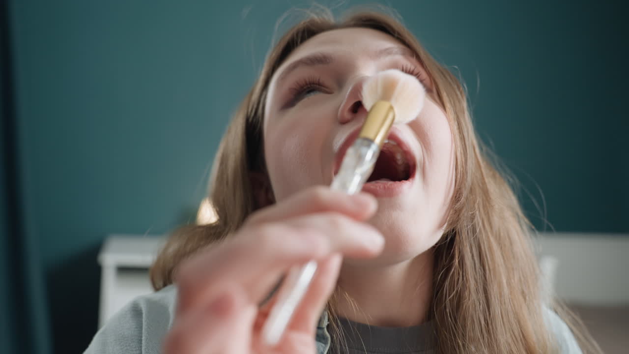Caucasian teen holding makeup brush playfully, dotted glitter falling, closeup expressions transitioning from curiosity to surprise to laughter, bedroom setting, soft teal wall, cozy lamp background
