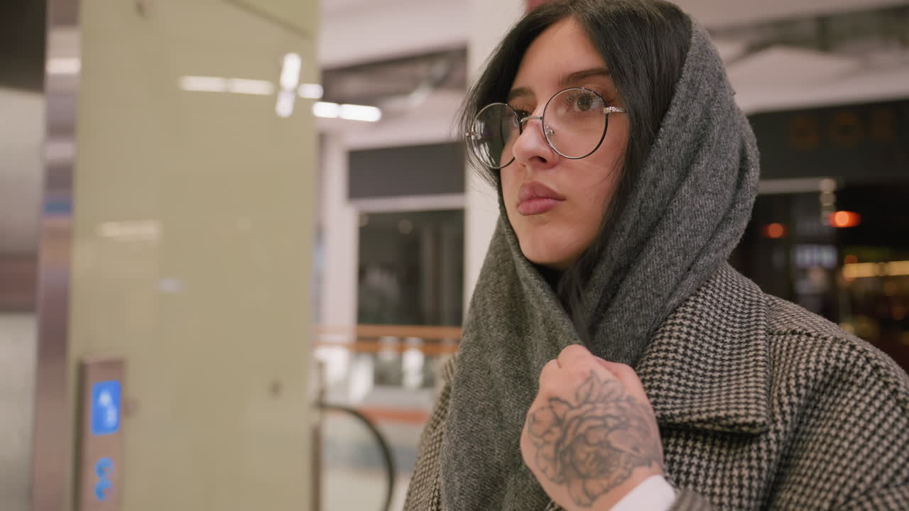 woman wearing glasses and scarf adjusts head covering while turning inside well-lit mall environment with modern interior and blurred background details in view
