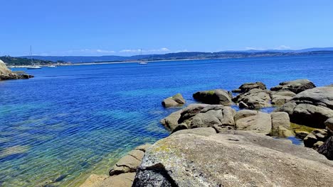 Felsiges-Ufer-Mit-Klarem-Wasser-Und-Segelbooten-Mit-Land-Am-Horizont