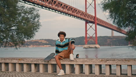 young man sitting skateboard at water view portrait. skater guy looking camera